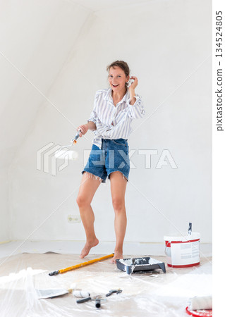 Portrait young adult beautiful single happy woman painting wall paint roller brush white color living room moving new home flat . Female painter decorating interior renovation residential apartment Portrait young adult beautiful single happy woman painting wall paint roller brush white color living room moving new home flat . Female painter decorating interior renovation residential apartment 134524805