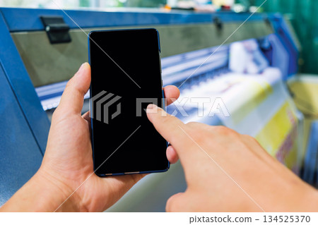 Hand holding smartphone with blank screen on large format printer printing large scale paper Hand holding smartphone with blank screen on large format printer printing large scale paper 134525370
