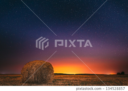 Night Starry Sky Above Field Meadow With Hay Bale After Harvest. Glowing Stars And Sunset Sunrise Lights Above Rural Landscape In August Month. Agricultural Colorful Background Copy Space Night Starry Sky Above Field Meadow With Hay Bale After Harvest. Glowing Stars And Sunset Sunrise Lights Above Rural Landscape In August Month. Agricultural Colorful Background Copy Space 134528857