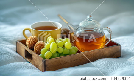 Breakfast tray with hot tea in a glass teapot and yellow cup, fresh green grapes, and dried longan fruit on a white bed Breakfast tray with hot tea in a glass teapot and yellow cup, fresh green grapes, and dried longan fruit on a white bed 134529305