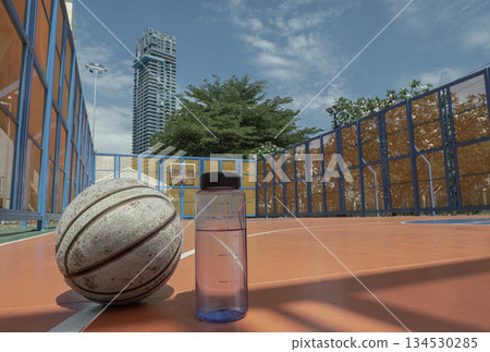 The bright blue sky and soaring modern building stand in stark relief to the grounded, worn basketball and the unassuming water bottle, all resting quietly on the vibrant orange court. 134530285