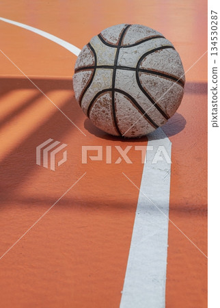 A battered basketball rests against the stark white leading lines of the vivid orange court. 134530287