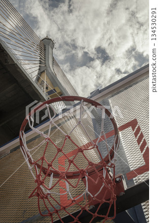 Gaze is drawn upward through the crimson and white basketball net to the industrial Ring Road Bridge and the dramatic sky beyond. 134530291