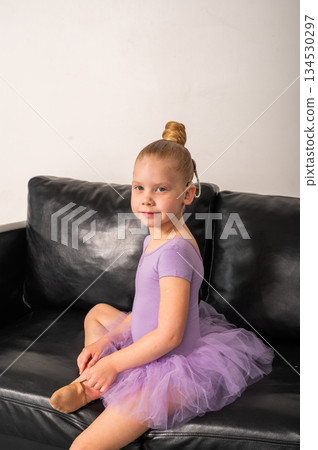 Little ballerina with cochlear implant sitting on black sofa and smiling. Inclusion, hearing awareness, and emotional resilience through confidence and art. 134530297
