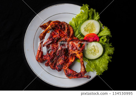 Ayam Taliwang, traditional grilled chicken from Lombok, Indonesia, served on a white plate with fresh salad, top view on black background. 134530995