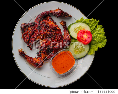 Ayam Taliwang, traditional spicy grilled chicken from Lombok, Indonesia, served on a white plate with fresh vegetable garnish and sambal chili sauce. Isolated on black background, top view. Ayam Taliwang, traditional spicy grilled chicken from Lombok, Indonesia, served on a white plate with fresh vegetable garnish and sambal chili sauce. Isolated on black background, top view. 134531000