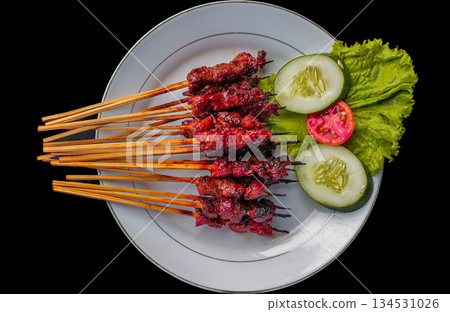Sate Rembiga, traditional spicy skewered beef or mutton meat from Lombok, Indonesia, served on white plate with fresh cucumber and tomato garnish on black background. Sate Rembiga, traditional spicy skewered beef or mutton meat from Lombok, Indonesia, served on white plate with fresh cucumber and tomato garnish on black background. 134531026