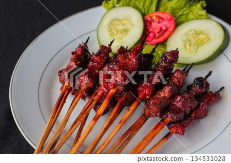 Sate Rembiga from Lombok, Indonesia. Traditional Indonesian spicy beef satay skewers served on a white plate with fresh vegetables. 134531028