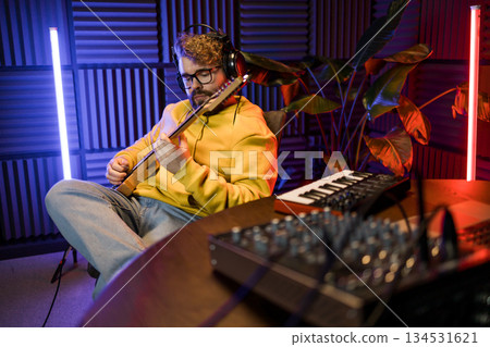 A focused male musician fine-tunes a guitar against the backdrop of a digital sound editing setup in a contemporary recording studio environment. A focused male musician fine-tunes a guitar against the backdrop of a digital sound editing setup in a contemporary recording studio environment. 134531621