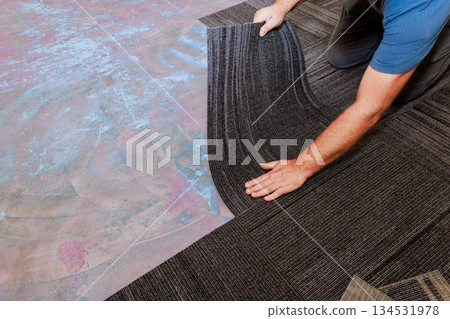 Worker carefully lays down carpet tiles on neat installation in new room 134531978