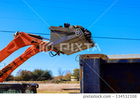 An excavator lifts bucket of soil to fill dumpster at busy construction site 134532108