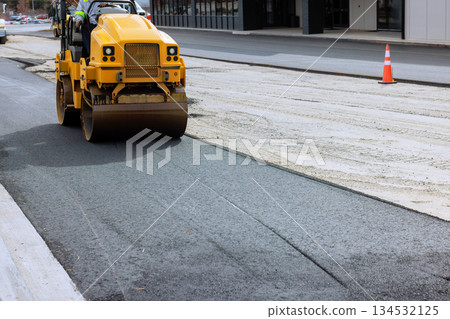 Steamroller roller compacts freshly laid asphalt on street under construction in bustling city setting. 134532125