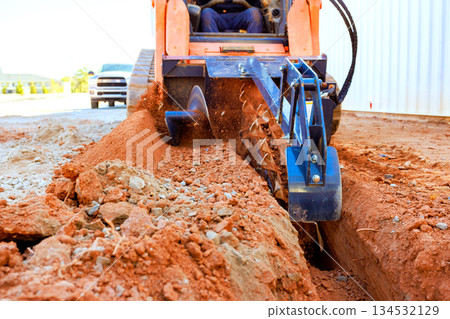 Worker operates trenching machine to create trench in dirt at construction site. 134532129