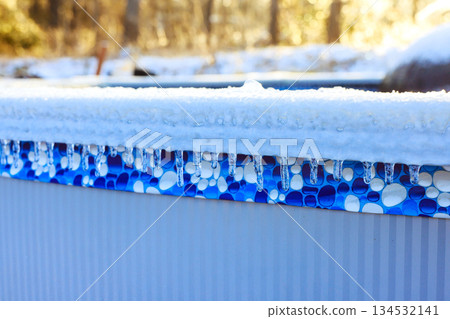 Frozen icicles drip from above ground pool edge amidst peaceful winter scene with trees snow. Frozen icicles drip from above ground pool edge amidst peaceful winter scene with trees snow. 134532141