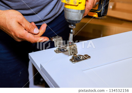 Carpenter uses power drill to attach hinges to cabinet in workshop setting. 134532156