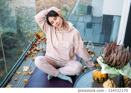 Woman stretches on yoga mat among autumn leaves on balcony. Holistic healing, emotional resilience, self-care rituals, stress management. 134532170