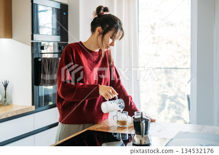 Woman pours frothed milk into glass mugs on kitchen counter during morning routine. Home barista skills, manual brewing, coffee aesthetics. Woman pours frothed milk into glass mugs on kitchen counter during morning routine. Home barista skills, manual brewing, coffee aesthetics. 134532176