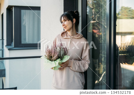 Woman holds potted heather plant on urban balcony. Urban sanctuary, mental restoration, residential wellness, greenery at home. 134532178
