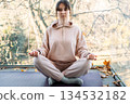 Woman sits with eyes closed in meditative pose on balcony during autumn. Self-care habits, home wellness sanctuary, tranquil lifestyle. 134532182