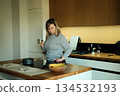 Woman stands in modern kitchen holding jar of spices while looking away from camera. Kitchen workflow, pantry organization, spice storage solutions. 134532193