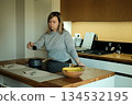 Woman stands in modern kitchen pouring seasoning from shaker into small pot on stove. Domestic mindfulness, cooking as meditation, evening wind-down, mental clarity, kitchen sanctuary. 134532195