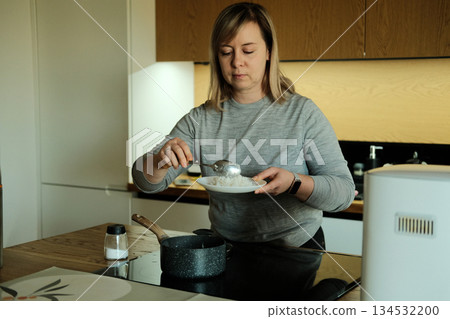 Woman serves cooked rice from pot onto plate in modern kitchen. Meal preparation, batch cooking, healthy eating habits, home wellness. Woman serves cooked rice from pot onto plate in modern kitchen. Meal preparation, batch cooking, healthy eating habits, home wellness. 134532200