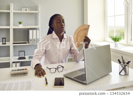 Young african american woman working at office using hand fan during heatwave in office. 134532634