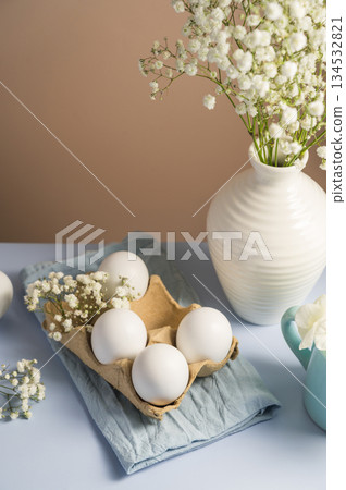 Easter eggs background. White eggs in cardboard box and gypsophila flowers in a vase. Spring holiday Easter eggs background. White eggs in cardboard box and gypsophila flowers in a vase. Spring holiday 134532821