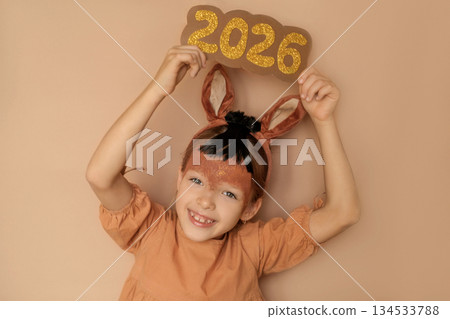 A laughing mischievous girl holds the golden numbers 2026 high above her, demonstrating the approach of the new year. A child in a costume and with horse makeup 134533788