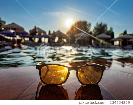 Sunlit summer poolside scene with stylish sunglasses resting on shimmering edge during golden hour vibes Sunlit summer poolside scene with stylish sunglasses resting on shimmering edge during golden hour vibes 134534239