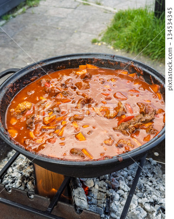 Communal outdoor feast featuring robust stew of meat and peppers slow cooked in heavy pot over charcoal flame 134534345
