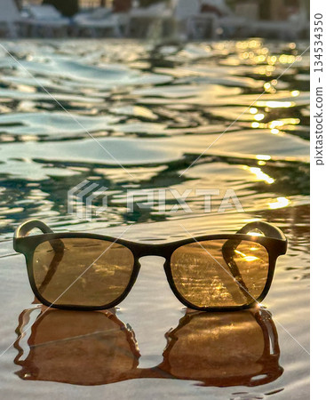 Stylish sunglasses reflecting shimmering golden ripples under warm golden hour glow on tranquil water surface 134534350