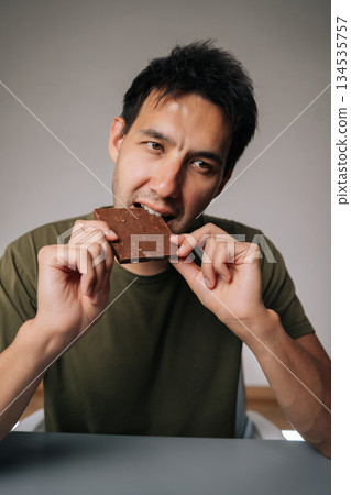 Vertical shot of happy Caucasian man sitting at table biting chocolate, savoring rich flavor of dark chocolate bar, blissful enjoyment as takes satisfying and delicious bite, on isolated background. 134535757