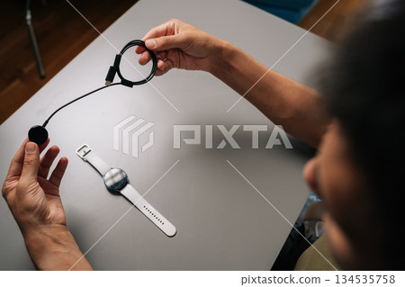 High-angle view of from shoulder of young adult man holding smartwatch inductive charger, preparing to power up smart watch, sitting at table. Concept of healthy lifestyle. 134535758