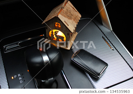 Toy house wooden figurine on the car center panel near the gear knob and car key. Conceptual stock photo on the topic of Christmas present  134535963