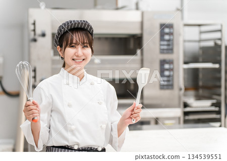 A young Asian woman (pastry chef) wearing a chef's coat working in a confectionery workshop, cake shop kitchen, or bakery A young Asian woman (pastry chef) wearing a chef's coat working in a confectionery workshop, cake shop kitchen, or bakery 134539551