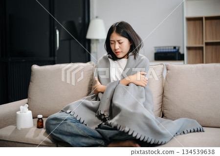 Asian woman suffering from flu symptoms, covered with a blanket on a sofa. Concept of illness, healthcare, and recovery. 134539638