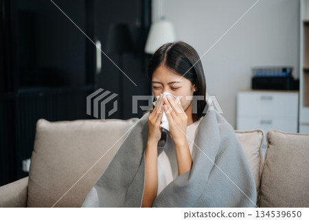 Asian woman suffering from flu symptoms, covered with a blanket on a sofa. Concept of illness, healthcare, and recovery. 134539650