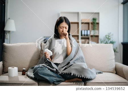Asian woman suffering from flu symptoms, covered with a blanket on a sofa. Concept of illness, healthcare, and recovery. 134539652
