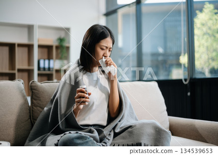 Asian woman suffering from flu symptoms, covered with a blanket on a sofa. Concept of illness, healthcare, and recovery. 134539653