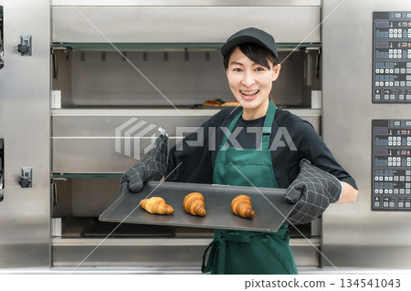 一名女廚房工作人員正在從烤箱裡取出麵包和羊角麵包。 一名女廚房工作人員正在從烤箱裡取出麵包和羊角麵包。 134541043