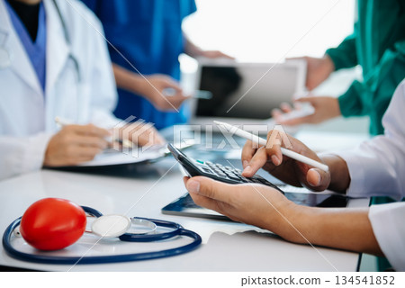 Medical team having a meeting with doctors in white lab coats and surgical scrubs seated at a table discussing a patients working online using computers Medical team having a meeting with doctors in white lab coats and surgical scrubs seated at a table discussing a patients working online using computers 134541852
