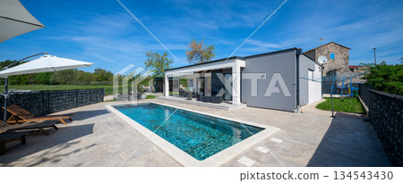 Modern modular house backyard featuring a rectangular swimming pool, large patio with wooden sun loungers, and umbrellas against a blue sky. Contemporary prefabricated home design 134543430