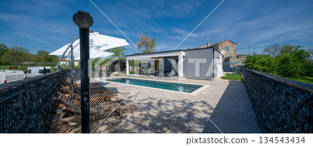 Modern modular house backyard featuring a rectangular swimming pool, large patio with wooden sun loungers, and umbrellas against a blue sky. Contemporary prefabricated home design Modern modular house backyard featuring a rectangular swimming pool, large patio with wooden sun loungers, and umbrellas against a blue sky. Contemporary prefabricated home design 134543434