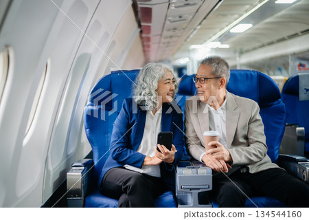 Senior Asian couple capturing memories on a plane, blending travel with technology. Smiling, stylish, Senior Asian couple capturing memories on a plane, blending travel with technology. Smiling, stylish, 134544160