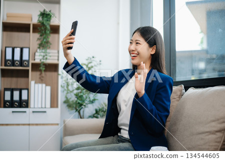 Asian woman, smiling and working at her desk in an office environment. She is wearing a business suit and holding documents while using a laptop. Asian woman, smiling and working at her desk in an office environment. She is wearing a business suit and holding documents while using a laptop. 134544605