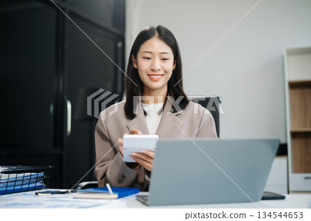 Asian woman, smiling and working at her desk in an office environment. She is wearing a business suit and holding documents while using a laptop. Asian woman, smiling and working at her desk in an office environment. She is wearing a business suit and holding documents while using a laptop. 134544653