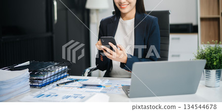 Young Asian woman, smiling and working at her desk in an office environment. She is wearing a business suit and holding documents Young Asian woman, smiling and working at her desk in an office environment. She is wearing a business suit and holding documents 134544678