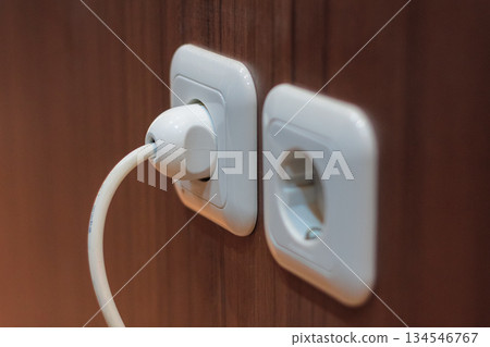 Closeup view of white power plug inserted into a European standard electrical socket on a wooden surface. Closeup view of white power plug inserted into a European standard electrical socket on a wooden surface. 134546767