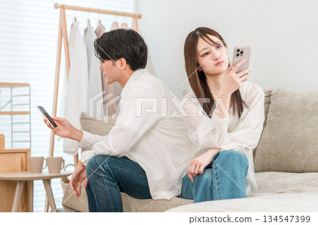 A couple sitting on the sofa in a tense atmosphere, looking at each other's smartphones, a married couple (couple quarrel, fight, misunderstanding) A couple sitting on the sofa in a tense atmosphere, looking at each other's smartphones, a married couple (couple quarrel, fight, misunderstanding) 134547399